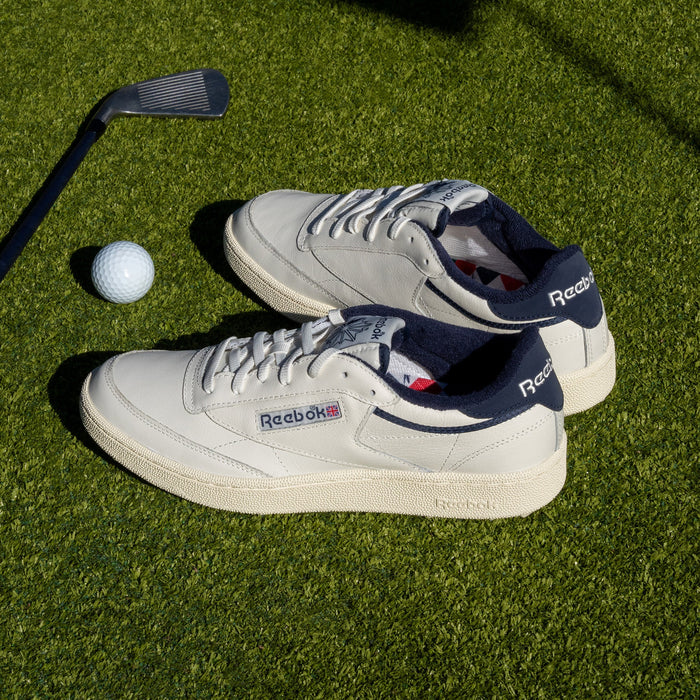 A pair of Reebok Men's Club C Golf Shoes in off-white with navy accents and a spikeless outsole sit on green grass next to a golf club and ball under bright sunlight.