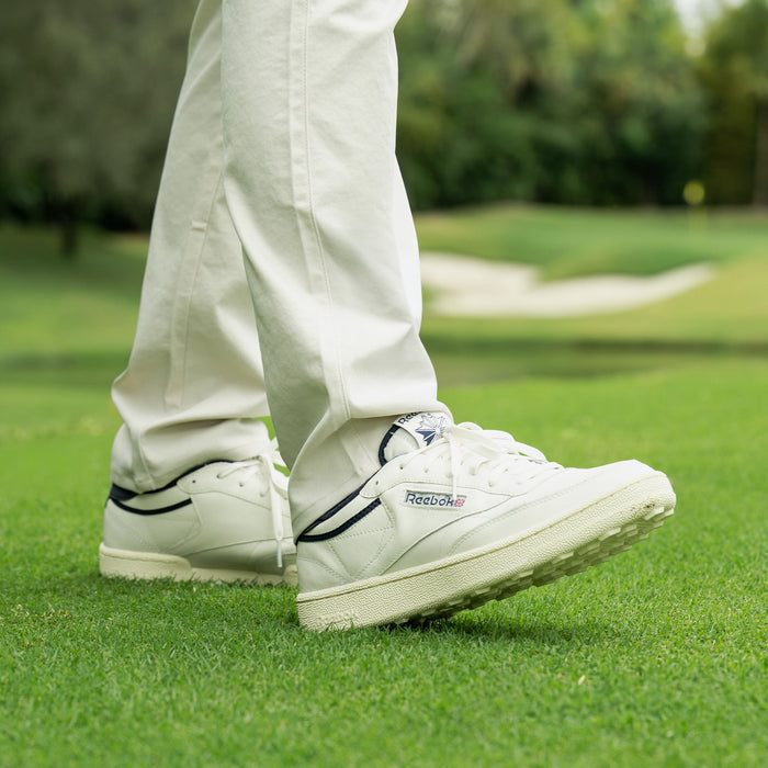 A person in white Reebok Men's Club C Golf Shoes with a spikeless outsole and white pants stands on green grass, with a golf course and trees blurred in the background.
