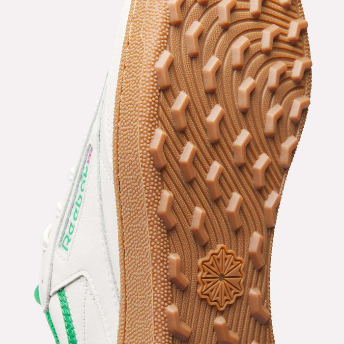Close-up of the sole on Reebok Men's Club C Golf Shoes. The gum-colored, spikeless outsole has a textured geometric tread and a central star-shaped emblem, with green accents highlighting the white sneaker.