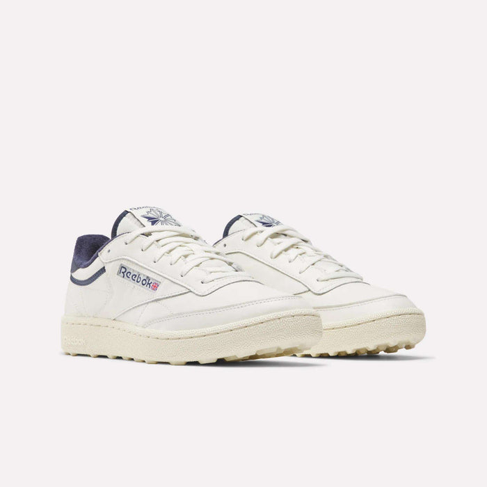 Men's Club C Golf Shoes by Reebok in off-white, featuring beige soles, navy accents on tongue and heel, side Reebok logo with Union Jack, and spikeless outsoles inspired by classic golf shoes.