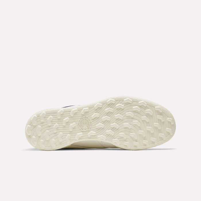 The image shows the off-white, textured sole of the Reebok Men's Club C Golf Shoes. It features a spikeless outsole with a patterned grip and starburst design near the center. The background is plain and light colored.