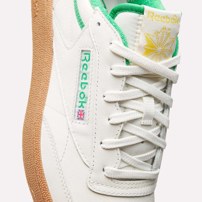 Close-up of Reebok Men's Club C Golf Shoes in white with green trim, featuring the green Reebok logo, British flag detail, gum rubber spikeless outsole, and a tongue with a yellow starburst and Reebok branding.