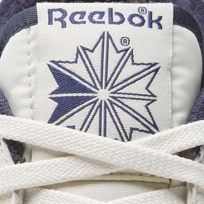 Close-up of the Men's Club C Golf Shoes by Reebok: a white tongue with blue Reebok logo and starburst, partially covered by laces. Crafted from water-resistant leather for lasting comfort on the course.
