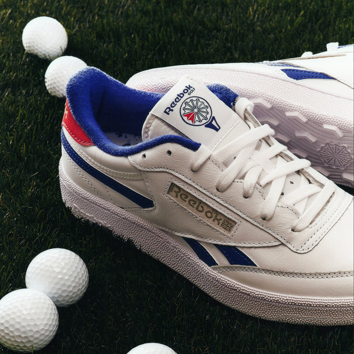 A pair of Reebok Men's Club C Revenge Golf Shoes with blue and red accents sit on green grass among white golf balls. These water-resistant shoes feature a spikeless outsole, combining style and performance for the course.