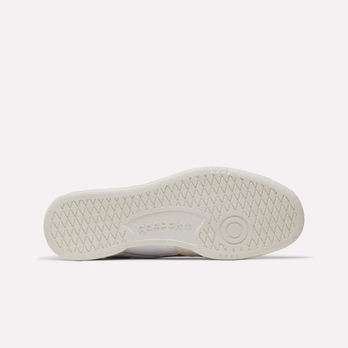 The image shows the bottom of a white Reebok Men's Club C Revenge Shoe sole, with a circular tread on the right, zigzag grip across the sole, and an embossed Reebok logo—highlighting its premium men’s design.
