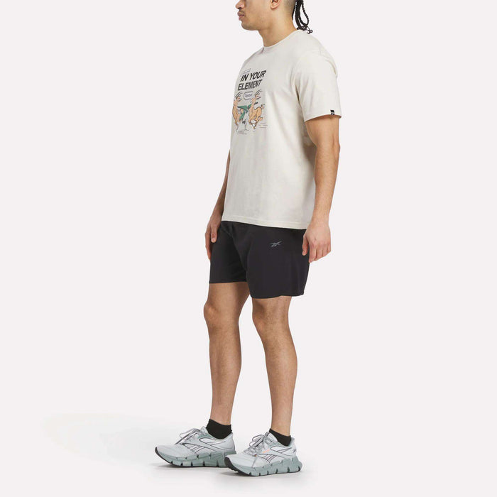 A person stands sideways against a plain white background wearing a Reebok Men's Club Graphic T-Shirt, black shorts, black socks, and light gray athletic sneakers.