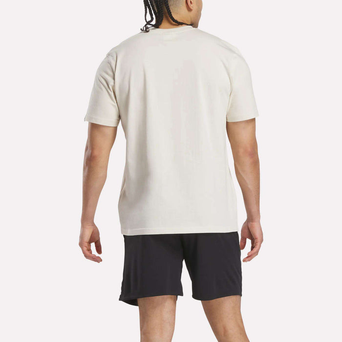 A man with braided hair faces away from the camera, wearing a Reebok Men's Club Graphic T-Shirt in plain white and black shorts, set against a light background.