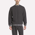 A man wears the Reebok Men's Club Sport Textured Jacket with matching pants, featuring white and gray striped cuffs and collar, standing against a plain light background.