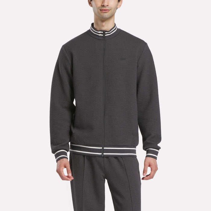 A man wears the Reebok Men's Club Sport Textured Jacket with matching pants, featuring white and gray striped cuffs and collar, standing against a plain light background.