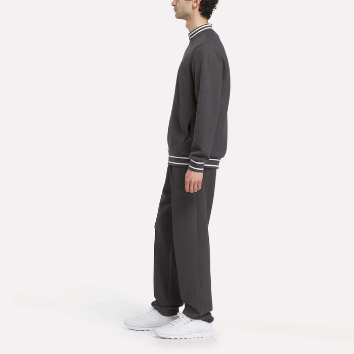 A man in profile wears the Reebok Men's Club Sport Textured Jacket in dark gray with white-striped cuffs and collar, paired with white sneakers, standing against a plain white background.