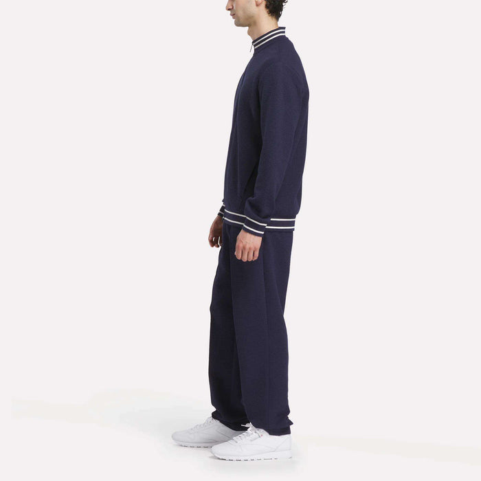 A man in profile wears a sporty navy tracksuit with white-striped cuffs and collar, featuring the Reebok Men's Club Sport Textured Jacket and white sneakers, standing against a plain white background.