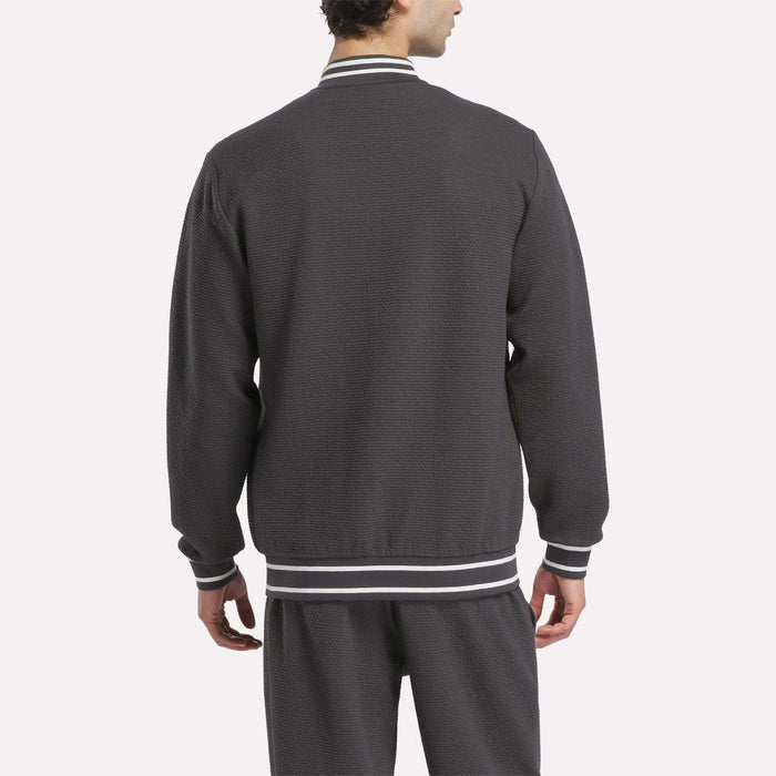 A man is seen from behind wearing the Reebok Men's Club Sport Textured Jacket in dark gray, featuring white and gray striped cuffs, collar, and waistband. Matching pants complete the sporty look against a plain white background.