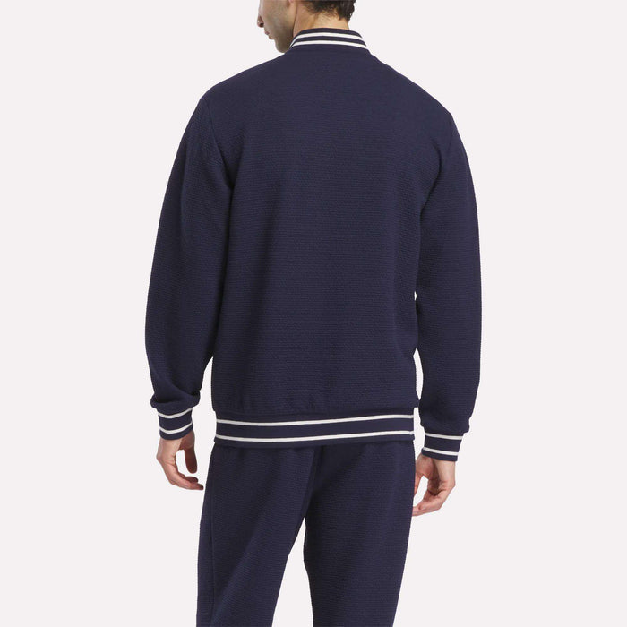 A man is seen from behind wearing the Reebok Men's Club Sport Textured Jacket in navy blue with white striped trim on the collar, cuffs, and waistband, standing against a plain white background.