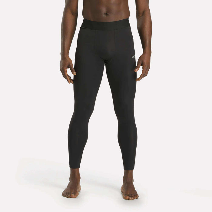 A person stands barefoot against a plain white background, wearing Reebok Men's Core Training Tights in black with a small white logo on the left thigh.