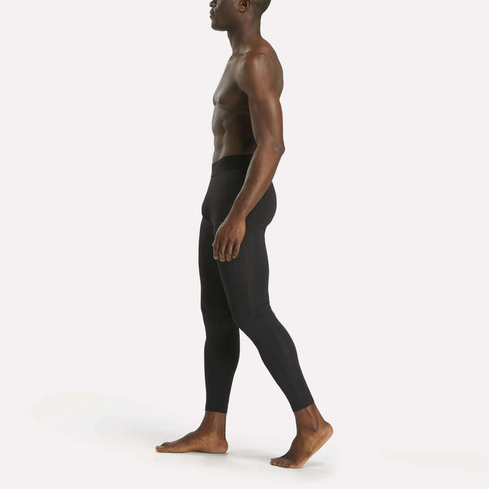 A shirtless man stands in profile, barefoot, wearing Reebok Men's Core Training Tights with a sleek fit and sweat-wicking performance, set against a plain white background.