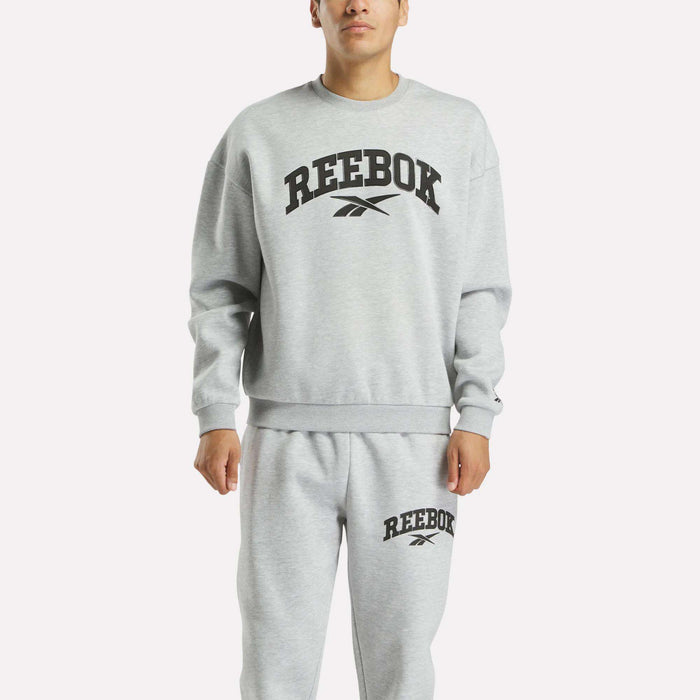 A man stands against a plain white background wearing a light gray Reebok Men's Cottle Sweatshirt and matching sweatpants, both displaying the black Reebok logo and text on the front.
