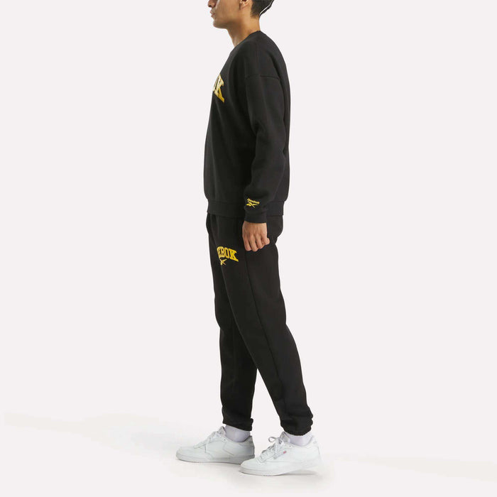 A person stands in profile wearing the Reebok Men's Cottle Sweatshirt and matching sweatpants with yellow text and logos, paired with white sneakers, against a plain light background.