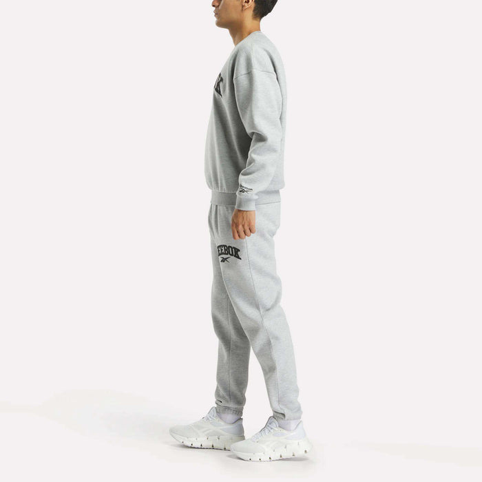 A person stands sideways in a matching grey Reebok Men's Cottle Sweatshirt and joggers with black logos, paired with white sneakers, against a plain white background.