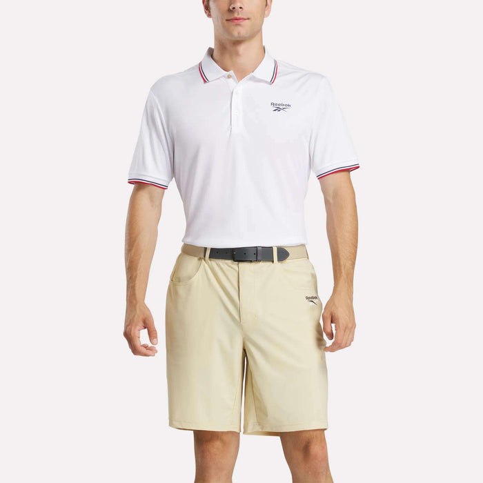 A man in a white Reebok Men's Country Club Oxford Pique Polo Shirt with red trim, beige shorts, and a black belt stands against a plain light background.