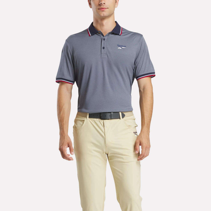 A man wearing a Reebok Men's Country Club Oxford Pique Polo Shirt stands facing forward with his arms relaxed by his sides, against a plain background.