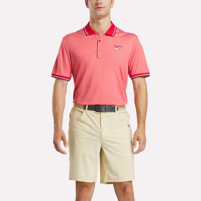 A man faces forward wearing a pink Reebok Men's Country Club Oxford Pique Polo Shirt with white and red trim on the collar and sleeves, an embroidered logo on the left chest, beige shorts, and a black belt.