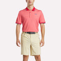 A man faces forward wearing a pink Reebok Men's Country Club Oxford Pique Polo Shirt with white and red trim on the collar and sleeves, an embroidered logo on the left chest, beige shorts, and a black belt.