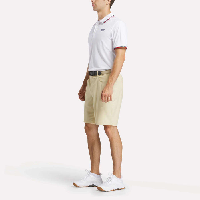 A man stands against a plain light background wearing a Reebok Men's Country Club Oxford Pique Polo Shirt, featuring red trim, paired with beige knee-length shorts, a black belt, and white sneakers.
