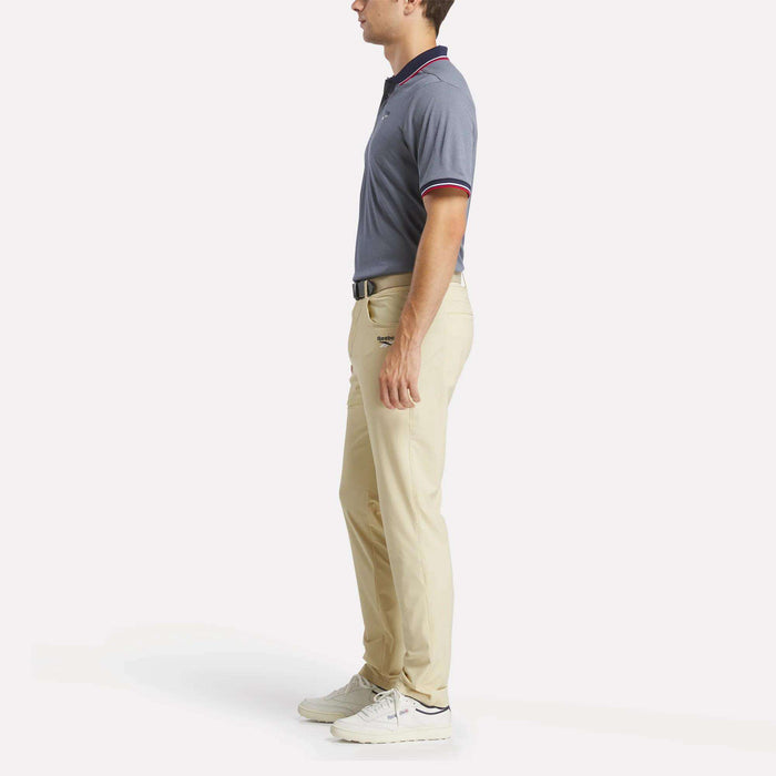 A man stands in profile wearing a men's Reebok Country Club Oxford Pique Polo Shirt, beige pants, white sneakers, and a black belt against a plain white background.