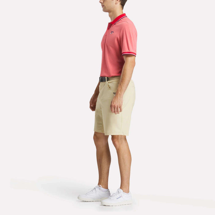 A man stands in profile against a plain white background wearing a red Men's Country Club Oxford Pique Polo Shirt by Reebok, beige shorts, a black belt, and white sneakers.