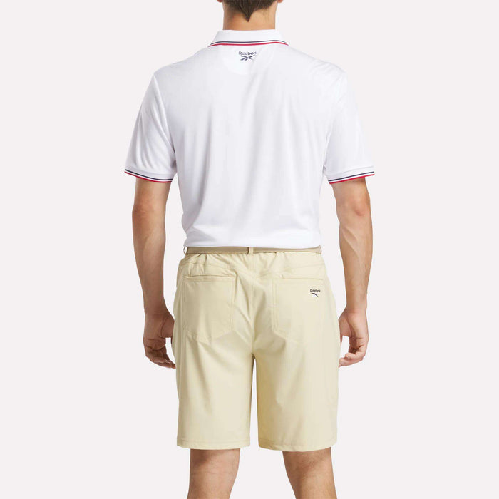 A man faces away wearing a white Reebok Men's Country Club Oxford Pique Polo Shirt with red trim and tan shorts, set against a plain light background.