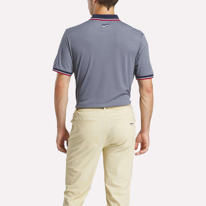 A man, shown from the back, wears a gray Reebok Men's Country Club Oxford Pique Polo Shirt with red and white trim and light beige pants, standing against a plain white background.