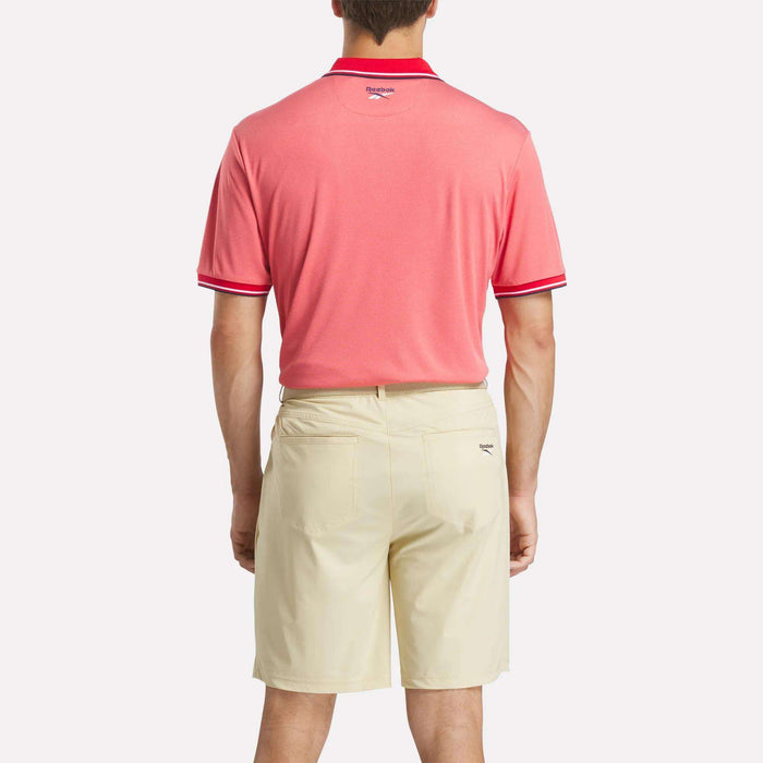A man stands with his back to the camera wearing a pink Reebok Men's Country Club Oxford Pique Polo Shirt with red trim and light beige shorts, set against a plain white background.