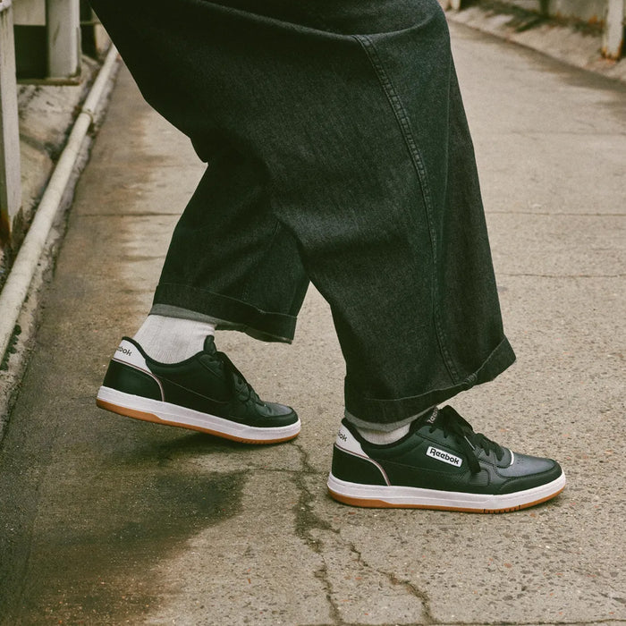 A person stands on a sidewalk, wearing black Reebok Men’s Court LXS Shoes with white soles and white socks. Only their lower legs and the wide-legged dark jeans are visible. These Reebok shoes are crafted from premium full grain leather, offering a classic and durable style.