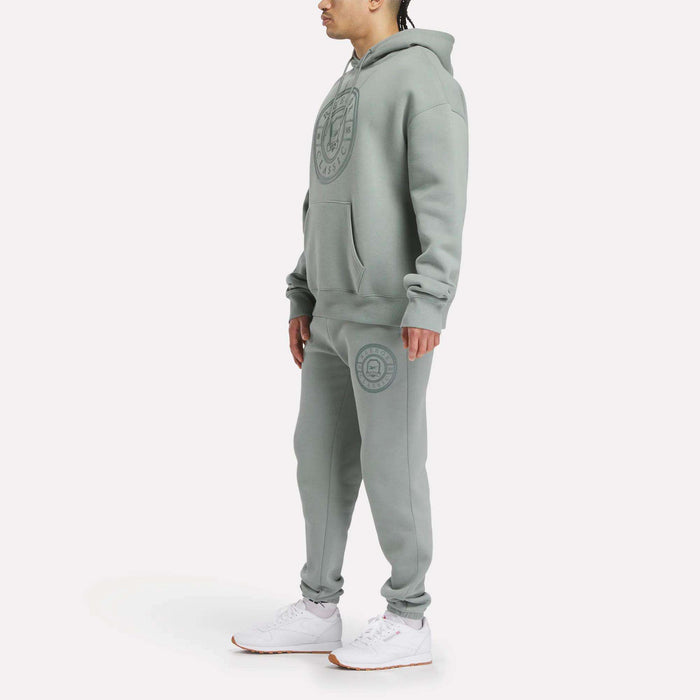 A person stands wearing a relaxed fit Reebok Men's Crest Hoodie and sweatpants set in light gray fleece with large circular logos, paired with white sneakers, against a plain white background.