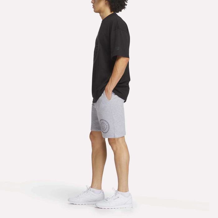A person stands in profile with hands in pockets, wearing a black oversized t-shirt, Reebok Men's Crest Shorts with a circular design, and white sneakers against a plain white background.