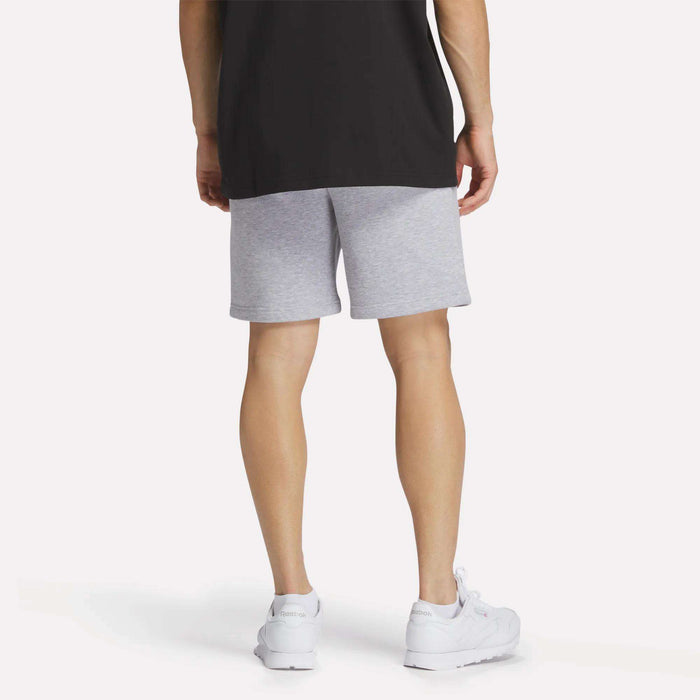 A person wearing a black T-shirt, Reebok Men's Crest Shorts in light gray (relaxed fit), and white sneakers is shown standing with their back to the camera against a plain white background.