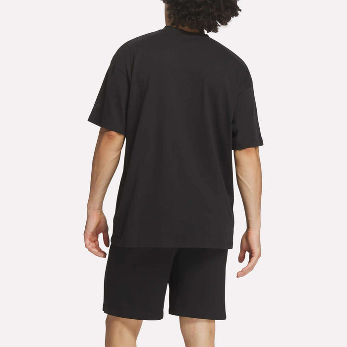 A person with curly hair is shown from the back against a plain white background, wearing a loose black Reebok Men's Crest T-Shirt and black shorts for everyday comfort.