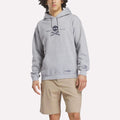 Wearing a light gray Reebok Men's Crushers Hoodie featuring a skull and crossed golf clubs graphic, paired with tan shorts, a person stands against a plain background.