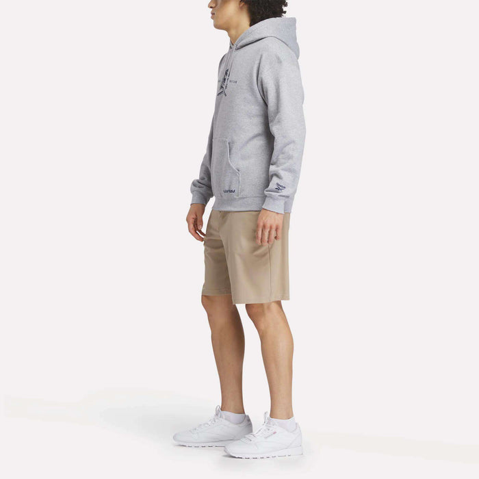 A person stands sideways in a Men’s Crushers Hoodie by Reebok, paired with tan shorts and white Reebok sneakers, against a plain white background.