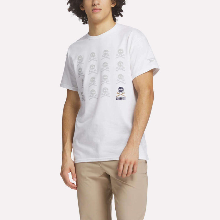 A man wears the Reebok Men's Crushers Icon Tee, featuring repeated gray skull and crossbones graphics on white, along with beige pants, standing against a plain light background.