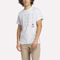 A man wears the Reebok Men's Crushers Icon Tee, featuring repeated gray skull and crossbones graphics on white, along with beige pants, standing against a plain light background.