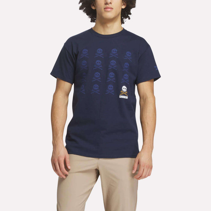A person wearing the Reebok Men's Crushers Icon Tee in navy blue with a repeating skull and crossbones pattern and tan pants, standing against a plain light background.