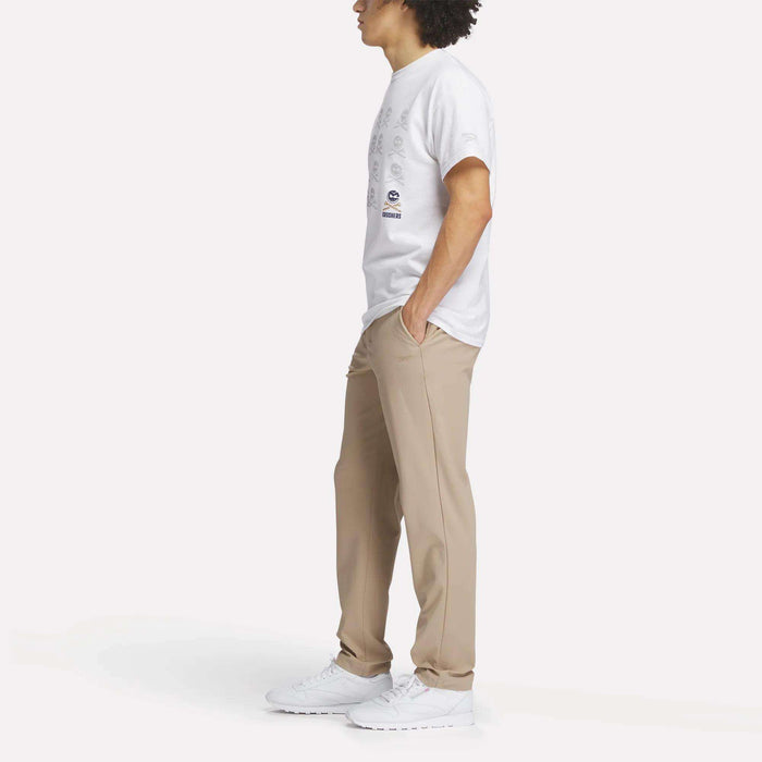 A person stands in profile with hands in pockets, wearing a white Reebok Men's Crushers Icon Tee, beige pants, and white sneakers against a plain white background.