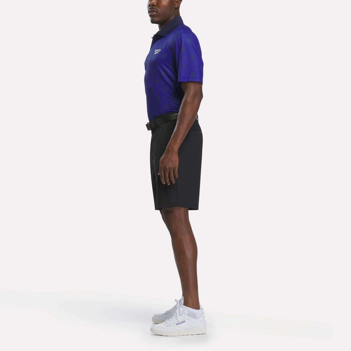 A man stands in profile wearing a dark blue Reebok Men's Crushers Polo Shirt, black shorts, white sneakers, and a black belt against a plain white background.