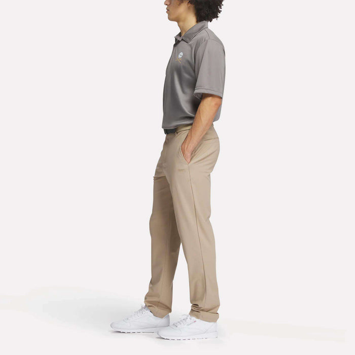 A person stands in profile with hands in pockets, wearing a gray Reebok Men's Crushers Replica Polo, tan pants, and white sneakers against a plain white background.