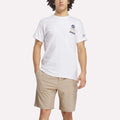 A person wears the Reebok Men's Crushers Stacked Logo Tee in white, featuring a small chest graphic, paired with light tan shorts, standing with one hand in their pocket against a plain white background.