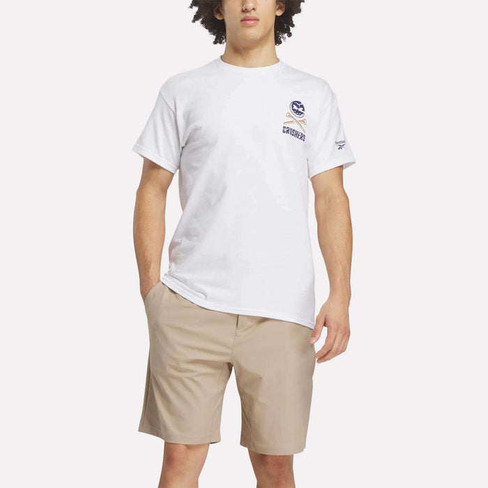 A person wears the Reebok Men's Crushers Stacked Logo Tee in white, featuring a small chest graphic, paired with light tan shorts, standing with one hand in their pocket against a plain white background.