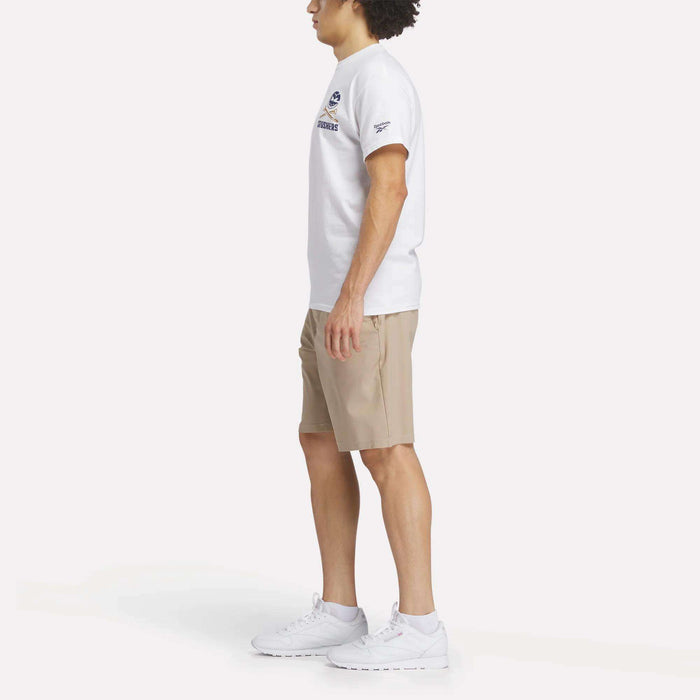 A man stands in profile against a plain white background wearing a Reebok Men's Crushers Stacked Logo Tee, beige shorts, and white Reebok sneakers.