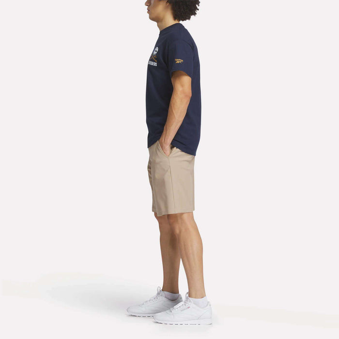 A person stands in profile wearing a navy Reebok Men's Crushers Stacked Logo Tee, beige shorts, and white sneakers, hands in pockets, against a plain white background.