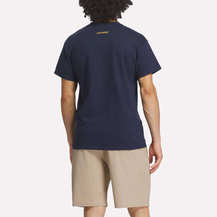 A person with curly hair is seen from behind, wearing the Reebok Men's Crushers Stacked Logo Tee in navy blue with a small yellow logo near the collar and light beige shorts, standing against a plain white background.